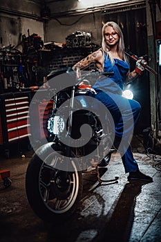 Female tattooed mechanic in work overalls hold a big wrench and posing for a camera in garage