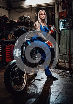 Female tattooed mechanic in work overalls hold a big wrench and posing for a camera in garage