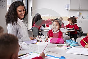 Female teacher and Chinese schoolgirl sitting at a table in an infant school class smiling to other kids, selective focus