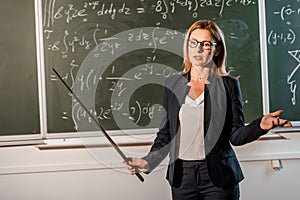 Female teacher in formal wear with wooden pointer explaining mathematical equations