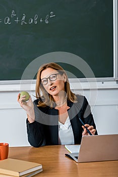 Female teacher in glasses sitting at computer desk and holding apple