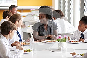 Female Teacher With Group Of High School Students Wearing Uniform Sitting Around Table And Eating Lunch In Cafeteria