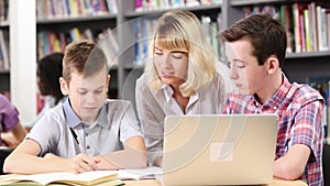 Female teacher helping two male high school students working at laptop