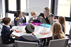 Female teacher kneeling to talk to a group of primary school kids sitting together at a round table eating their packed lunches