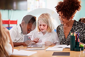 Female Teacher With Two Elementary School Pupils Wearing Uniform Using Digital Tablet At Desk