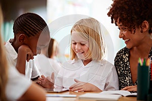 Female Teacher With Two Elementary School Pupils Wearing Uniform Using Digital Tablet At Desk