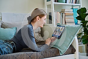 Female teenage student having group video conference, using digital tablet, lying on couch at home.