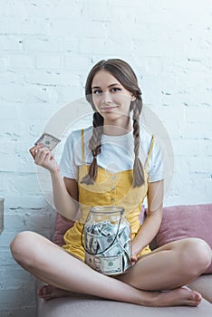 female teenager putting dollar banknote into glass jar for saving while sitting