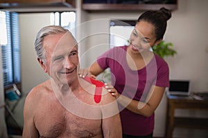 Female therapist applying elastic therapeutic tape on shoulder of smiling shirtless senior male pati