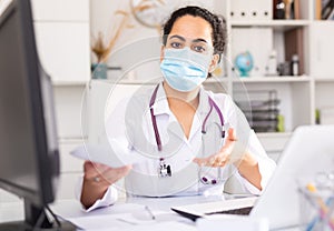 Female therapist in protective mask working at table with laptop in office