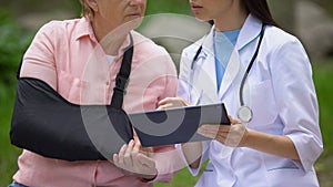 Female therapist showing tablet images to injured old lady in arm sling, rehab