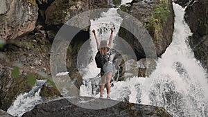 Female tourist with a backpack raises his hands up opposite a large waterfall standing on the edge of a cliff. Happy