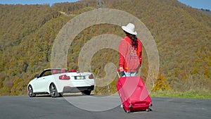 female tourist with big red suitcase walking to white cabriolet on road, rear view, travelling alone