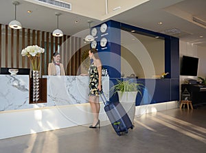 Female tourist with carry on luggage standing at hotel reception counter. Travelling, holiday and vacation concept