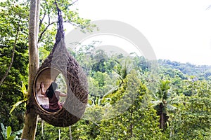 A female tourist is sitting on a large bird nest on a tree at Bali island