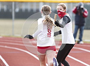 Female track runners exchanging the baton during a relay race wearing face masks