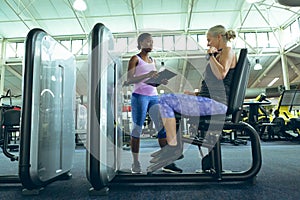 Female trainer assisting active senior woman to work out on leg curl machine