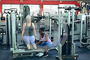 Female trainer assisting active senior woman to work out on leg curl machine