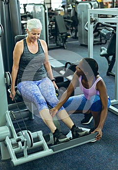 Female trainer assisting active senior woman to work out on leg curl machine