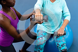 Female trainer assisting disabled senior woman to exercise with dumbbell in sports center