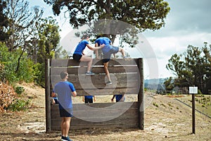 Female trainer assisting fit man to climb over wooden wall during obstacle course