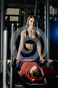 Female trainer helping guy to train in the gym Bar Bench Press.