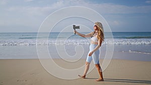 Female travel influencer records on pro camera with wireless lav mic on sandy beach. Curly-haired woman creates content
