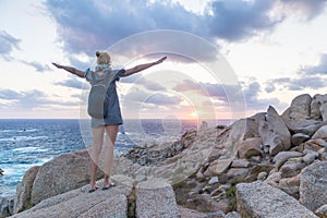 Female traveler, arms rised to the sky, watches a beautiful sunset on spectacular rocks of Capo Testa, Sardinia, Italy.