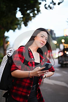 Female traveler with backpack using application of taxi service on smart phone while standing in city street.