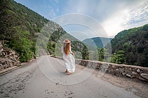 Female traveler in brown hat and white dress looking at amazing mountains and forest, wanderlust travel concept
