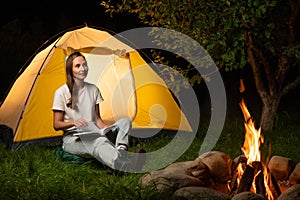 Female traveler is camping at a campsite. A young woman is sitting by the tent and reading a book in the camp by the