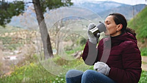 Female traveler drinks hot tea from thermos, rests sitting on a log bench among early spring forest