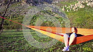 Female traveler having rest in orange hammock at mountain scenery.