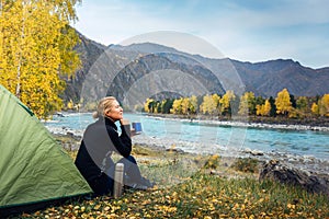 Female traveler sitting on grass near the tent, drinking coffee from thermos and admiring the beautiful view of river and