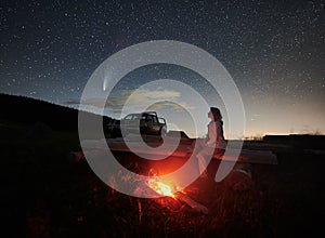 Female traveler sitting near campfire under night starry sky.