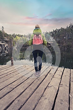 A female traveler is standing on a wooden pier near a beautiful lake in a pine forest.