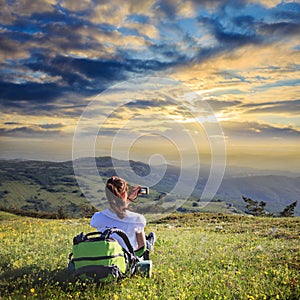 Female traveller with backpack making photo of mountain lanscape under sunset sky