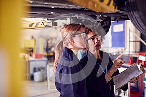 Female Tutor With Student Looking Underneath Car On Hydraulic Ramp On Auto Mechanic Course