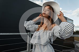 Female urban portrait of a stylish beautiful woman in summer clothes with a hat and a fashion striped top posing and looking at