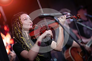 A female violinist plays the violin. Band performs on stage, rock music concert