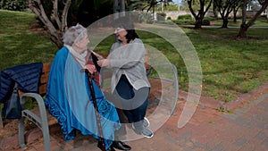 Female volunteer and elderly woman talk sitting on a bench in a city park