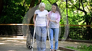 Female volunteer helping disabled senior woman walk with frame in park, support