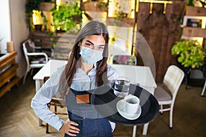 Female waitress in a medical protective mask serves the coffee in restaurant durin coronavirus pandemic representing new normal