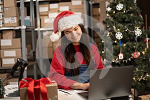 Female warehouse inventory manager using laptop computer and checking retail stock. Happy woman wear Santa hat working