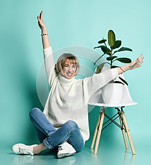 Female in white sweater, jeans, sneakers. Smiling, raised hands, sitting on floor. Chair, green ficus. Posing on blue background
