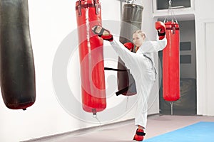 Female woking out karate raising leg and boxing heavy bag in gym.
