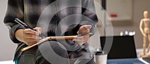 Female worker sitting on worktable and taking note on schedule book while looking on smartphone