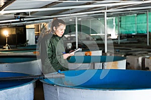 Female worker of trout farm watching fish and writing