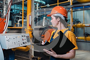 A female worker in uniform and helmet with a folder and a pen in her hand, writing meter readings. In the background-boiler room.