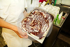 Female worker is working at ice cream factory is carrying creamy vanilla ice cream with chocolate dressing in steel container.